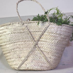 May include: A large, natural woven basket with a white, cream color. The basket has two handles and a woven X pattern across the front. The basket is filled with green leafy vegetables.