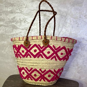 May include: A woven straw tote bag with pink embroidered geometric patterns and brown leather handles.