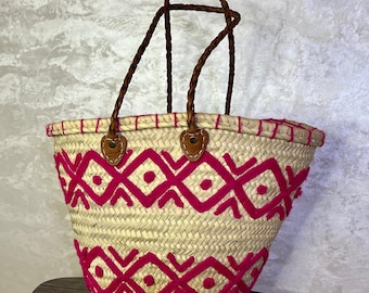 May include: A woven straw tote bag with pink embroidered geometric patterns and brown leather handles.