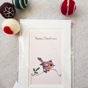 May include: A Christmas card featuring a watercolour illustration of a robin wearing a Santa hat, perched on a branch with holly. The card reads "Merry Christmas." Decorative felt balls in red, green, and white are scattered around the card.