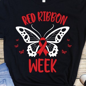 Könnte beinhalten: Schwarzes T-Shirt mit einem weißen Schmetterlingsdesign und einem roten Band. Der Text "Red Ribbon Week" ist auf dem Shirt aufgedruckt.