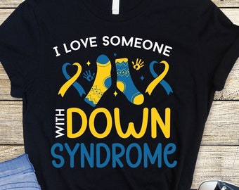 Pode incluir: Camiseta preta com o texto "Amo alguém com síndrome de Down" em letras amarelas e azuis. O design inclui duas meias amarelas e azuis com uma fita amarela e uma fita azul.
