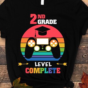 May include: Black t-shirt with a rainbow design and a video game controller. The text "2nd Grade Level Complete" is printed on the shirt.
