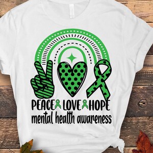Pode incluir: Camiseta branca com um sinal de paz, um coração e uma fita verde, preta e branca. O design está centralizado na camiseta e inclui o texto "Peace Love Hope mental health awareness".