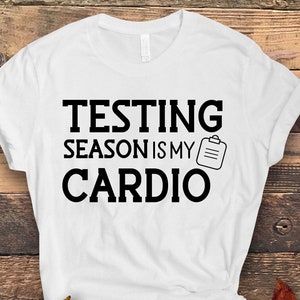 May include: White t-shirt with the text "TESTING SEASON IS MY CARDIO" in black, along with a small clipboard graphic. The shirt is displayed on a wooden background with autumn leaves.