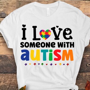 May include: White t-shirt with the text "i love someone with autism" in black. A colorful puzzle piece heart is in the middle of the text "love".