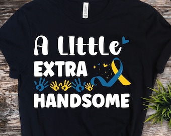 Pode incluir: Camiseta preta com texto branco que diz "A little extra handsome" com uma fita azul e amarela e impressões de mãos.