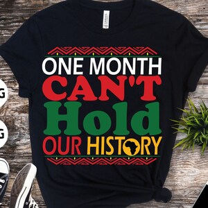 Puede incluir: Camiseta negra con un diseño gráfico que dice "One Month Can't Hold Our History" en letras rojas, verdes y amarillas. El gráfico también incluye una silueta negra del continente africano.
