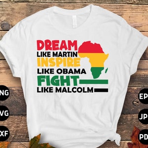 Può includere: Maglietta bianca con un design grafico che presenta una mappa stilizzata dell'Africa in rosso, giallo e verde. Il testo "DREAM LIKE MARTIN INSPIRE LIKE OBAMA FIGHT LIKE MALCOLM" è stampato in lettere nere sulla maglietta.