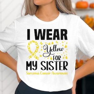 Op de afbeelding: Wit T-shirt met de tekst "Ik draag geel voor mijn zus" en een geel lint met vlinders. De tekst "Sarcoma Cancer Awareness" staat onder het lint.