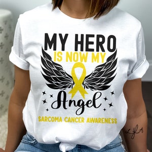 Op de afbeelding: Wit T-shirt met zwarte vleugels en een geel lint. De tekst op het shirt luidt "My Hero Is Now My Angel" en "Sarcoma Cancer Awareness".