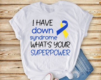 Pode incluir: Camiseta branca com o texto "I have down syndrome what's your superpower" em preto. Uma fita azul e amarela está à direita do texto.