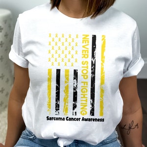 Op de afbeelding: Wit T-shirt met een verweerd Amerikaans vlagontwerp met gele linten en de tekst "NEVER STOP FIGHTING". De tekst "Sarcoma Cancer Awareness" is onder de vlag gedrukt.