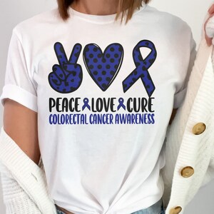 Puede incluir: Camiseta blanca con un signo de paz azul, un corazón y una cinta con lunares. El texto "Peace Love Cure Colorectal Cancer Awareness" está impreso debajo del diseño.