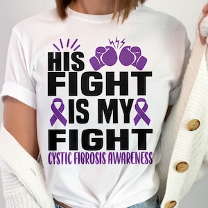Puede incluir: Camiseta blanca con una cinta morada y el texto "His Fight Is My Fight" y "Cystic Fibrosis Awareness".
