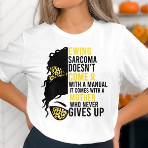 May include: White t-shirt with a black and yellow graphic design. The graphic features a woman's face with a yellow ribbon and the text "Ewing Sarcoma Doesn't Come & With a Manual It Comes With a Mother Who Never Gives Up".