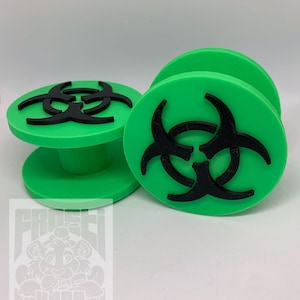 May include: Two green bottle stoppers featuring a black biohazard symbol. One stopper stands upright, the other lies on its side. The stoppers are made of a solid material and have a cylindrical base.