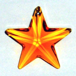 May include: A faceted, orange star-shaped crystal pendant with a hole at the top for hanging. The star has five points and a brilliant, light-reflecting surface. The crystal is a warm, amber color.