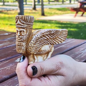 Può includere: Una piccola statua dettagliata di colore beige di un toro alato con testa umana, noto anche come lamassu, con una corona in testa. La statua è tenuta in una mano con smalto nero.
