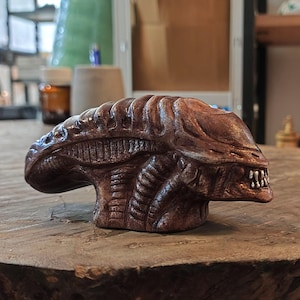 May include: A brown ceramic figurine of an alien creature with a detailed head and sharp teeth. The figurine is sitting on a wooden surface.