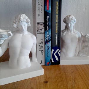 May include: White bookends shaped like a man holding a bowl and a woman holding a lyre. The bookends are on a wooden surface.