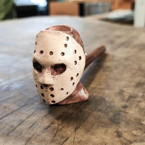 May include: A wooden pipe shaped like a hockey mask. The mask is white with black holes and a brown face.