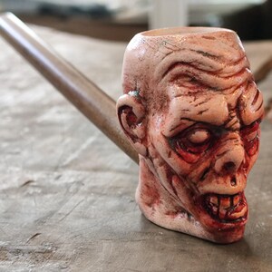 May include: A ceramic pipe shaped like a zombie head with a brown wooden stem. The head is painted with realistic details, including red blood splatters and a grotesque expression.