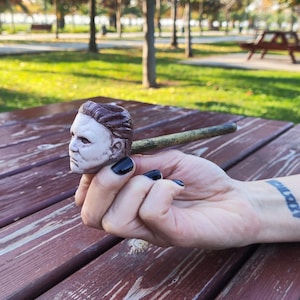 May include: A ceramic pipe shaped like the head of Michael Myers, the fictional character from the Halloween horror film franchise. The pipe is brown and white and has a black stem. The pipe is being held in a hand.