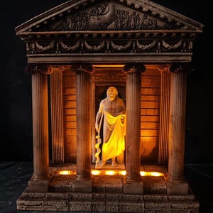 May include: A decorative temple structure featuring a central white statue of a bearded figure holding a staff. The architectural model has four columns, a pediment with relief carvings, and integrated warm yellow lighting illuminating the interior.