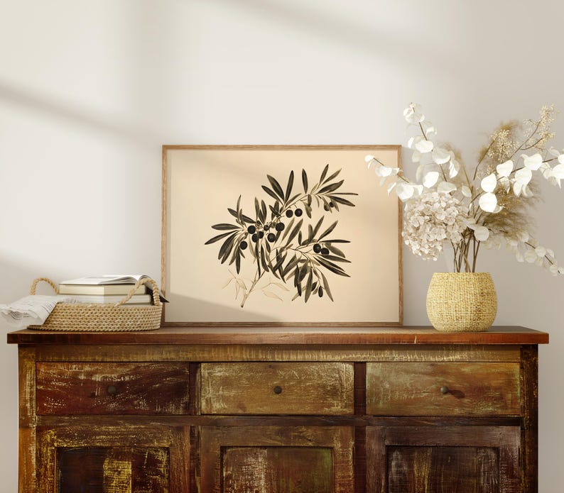 May include: A framed black and white botanical illustration of an olive branch with leaves and fruit. The frame is brown wood and the print is on a cream background. The print is sitting on a rustic wooden dresser with two drawers and two cabinet doors.