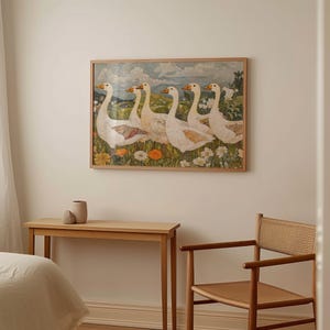 May include: A framed artwork depicts six white geese with orange beaks and accents, standing in a field of wildflowers. The artwork is in a light wood frame. A wooden table and chair are in the foreground.