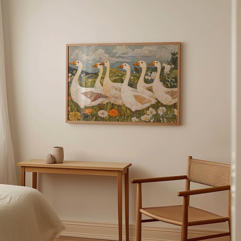 May include: A framed artwork depicts six white geese with orange beaks and accents, standing in a field of wildflowers. The artwork is in a light wood frame. A wooden table and chair are in the foreground.
