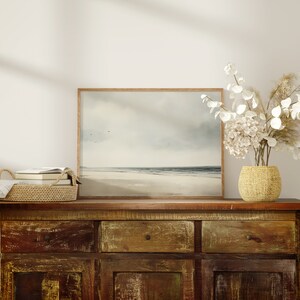 May include: A rustic wooden dresser with three drawers and two cabinet doors. A framed seascape painting hangs above the dresser. A woven basket filled with books sits on the dresser. A vase with white flowers sits on the dresser.