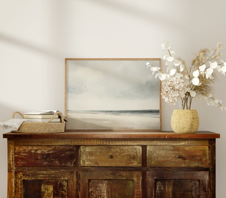 May include: A rustic wooden dresser with three drawers and two cabinet doors. A framed seascape painting hangs above the dresser. A woven basket filled with books sits on the dresser. A vase with white flowers sits on the dresser.