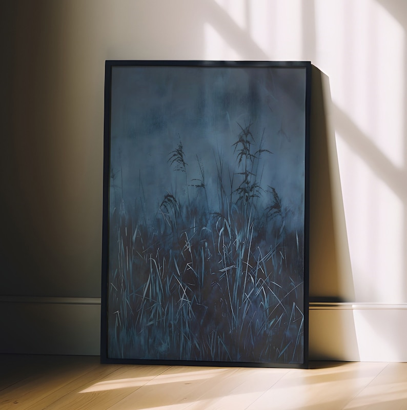 Puede incluir: Una impresi&oacute;n art&iacute;stica enmarcada que representa un paisaje oscuro y atmosf&eacute;rico de hierbas altas en tonos azules y grises. La obra de arte est&aacute; en un marco negro y se apoya contra una pared, con sombras de luz natural en el suelo de madera.