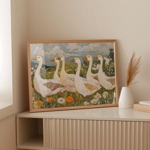 May include: A framed art print featuring a watercolor painting of six white geese with orange beaks and feet, standing in a field of wildflowers. The artwork has a light brown wooden frame and is displayed on a white cabinet.
