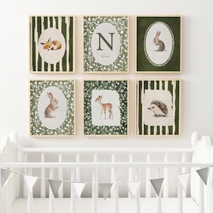May include: Six framed watercolor prints featuring woodland animals. The prints include a fox, a deer, a rabbit, a hedgehog, and an initial. Each print has a light wood frame and is set against a white background. The prints are above a white crib.