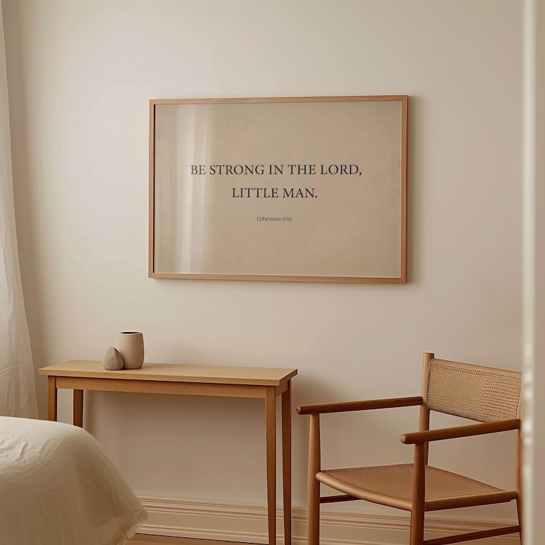 May include: A framed print with the text "BE STRONG IN THE LORD, LITTLE MAN. Ephesians 6:10" on a beige background. The print is hanging on a white wall above a wooden table and chair.