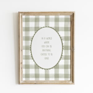May include: A framed print with a light brown wooden frame. The print features a green and white checkered pattern background. An oval shape in the center contains the text: "IN A WORLD WHERE YOU CAN BE ANYTHING, CHOOSE TO BE KIND."