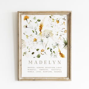 May include: Framed floral print featuring the name "Madelyn" in a serif font, with a watercolor design of yellow and white flowers and green leaves. Below the name are words like "Marvel" and "Adore". The frame is a light brown color.