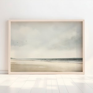 May include: A framed painting of a seascape with a cloudy sky and a sandy beach. The water is a light blue and the sand is a light brown. There are a few birds flying in the sky.