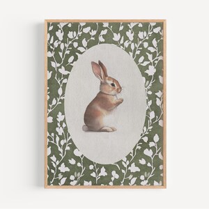 May include: A framed watercolor print of a brown and white rabbit. The rabbit is centered in an oval shape against a light green background. The oval is surrounded by a border of white floral designs, all within a wooden frame.