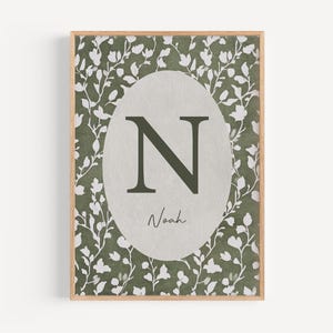May include: A framed print featuring a botanical design with a light brown wooden frame. The print has a green and white floral pattern background. A gray oval in the center displays the letter "N" in dark green, with the name "Noah" below.
