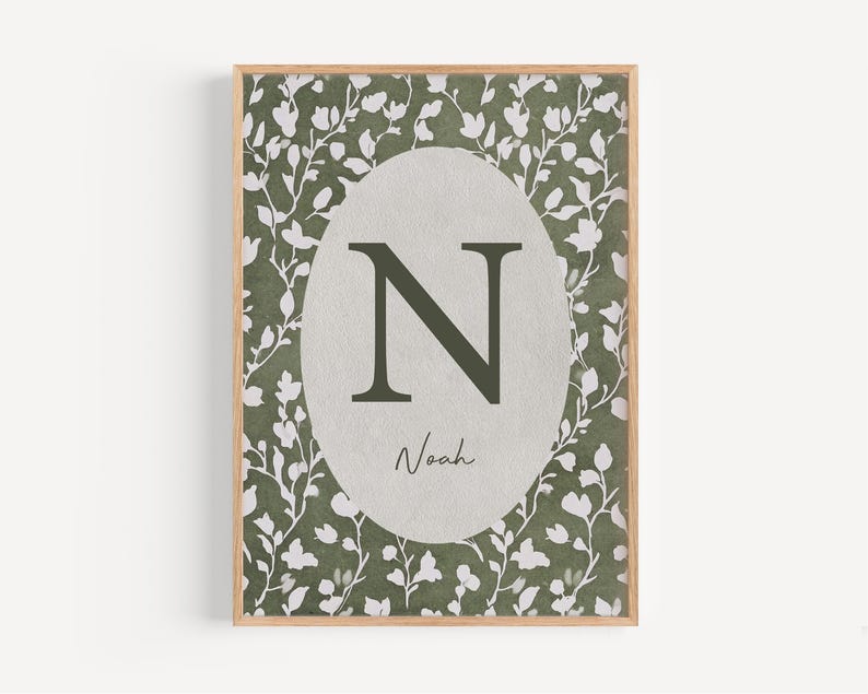 May include: A framed print featuring a botanical design with a light brown wooden frame. The print has a green and white floral pattern background. A gray oval in the center displays the letter "N" in dark green, with the name "Noah" below.