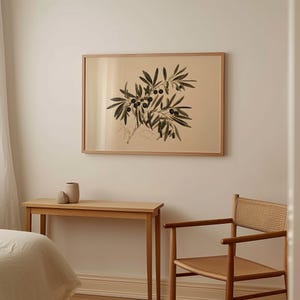 May include: A framed print of a black and white illustration of an olive branch with leaves and olives. The print is hanging on a white wall above a wooden table and chair.