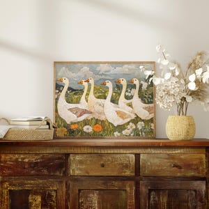 May include: A framed artwork featuring six white geese with orange beaks and feet, standing in a field of wildflowers. The artwork is displayed on a rustic wooden cabinet, with a woven basket and vase of dried flowers.