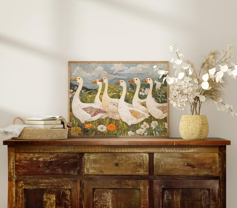 May include: A framed artwork featuring six white geese with orange beaks and feet, standing in a field of wildflowers. The artwork is displayed on a rustic wooden cabinet, with a woven basket and vase of dried flowers.