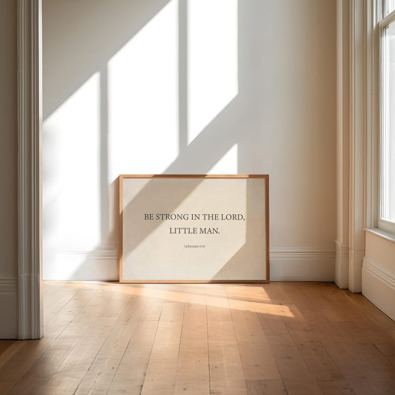 May include: A framed print with a beige background and a wooden frame. The print features the text "Be strong in the Lord, little man." in black font.