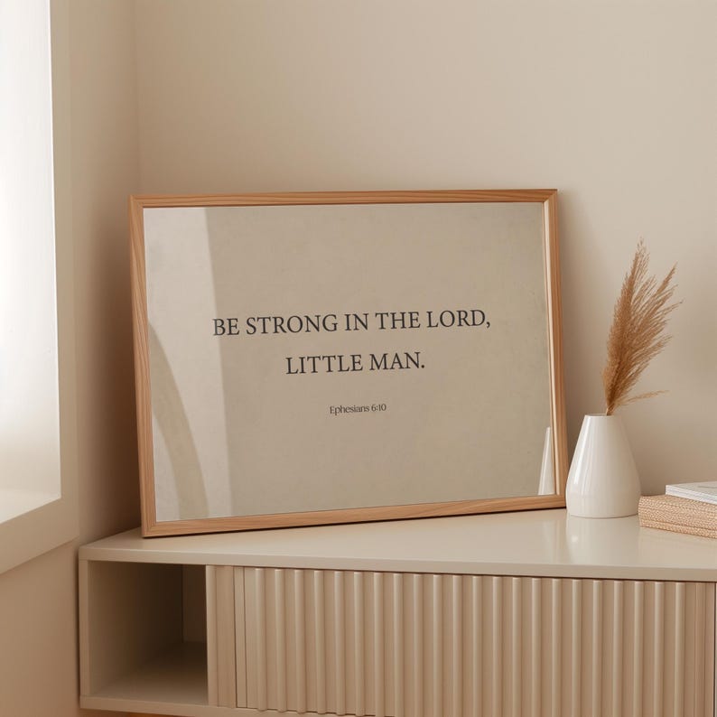 May include: A framed print with a beige background and a black inscription that reads "Be strong in the Lord, little man. Ephesians 6:10". The frame is made of light wood and is sitting on a light beige dresser with a ribbed front.
