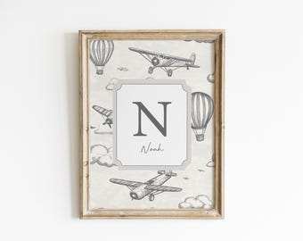 Puede incluir: Lámina enmarcada con temática de aviación vintage. La obra de arte incluye ilustraciones de aviones, globos aerostáticos y nubes en tonos grises. Un marco central muestra la letra "N" y el nombre "Noah". El marco tiene un aspecto de madera envejecida.